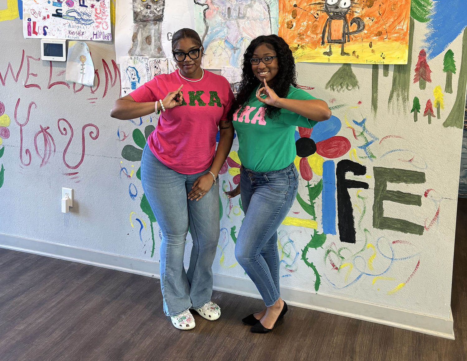 Image of two members  of AKA standing in front of a collection of children's paintings wearing Alpha Kappa Alpha shirts