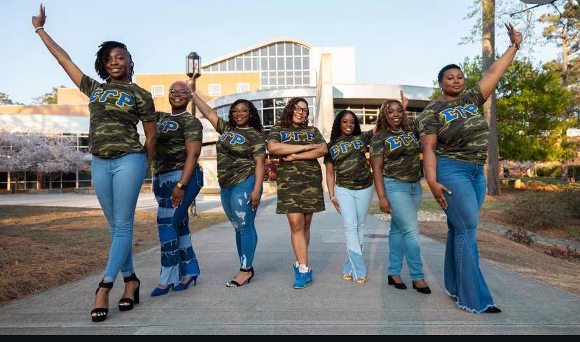 Image of members of Rho Lambda Chapter of Sigma Gamma Rho standing in front of the University Center, wearing blue and gold Greek Lettered paraphenalia over camoflauged clothing and jeans.