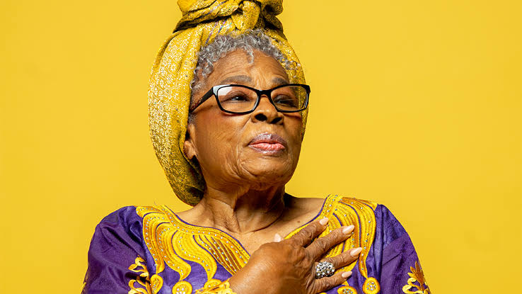 A portrait of Opal Lee, known as the 'Grandmother of Juneteenth.' She is wearing a vibrant purple and yellow traditional outfit with intricate embroidery and a matching head wrap. Her glasses frame her face, and she is holding one hand over her chest in a gesture that conveys pride and dignity. The background is a bright yellow, which complements her outfit.